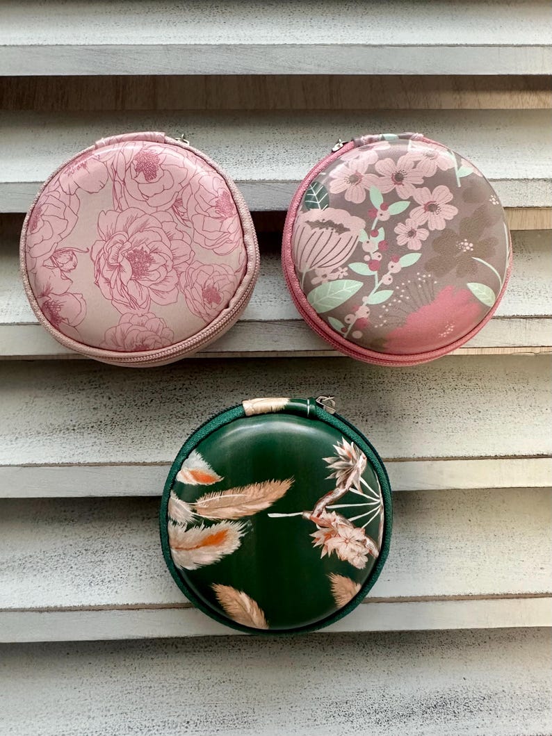 May include: Three round, zippered cases with floral and feather designs. The top case is light pink with a peony pattern. The middle case has a brown background with pink flowers. The bottom case is dark green with feather and floral accents.