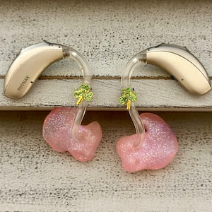 May include: Two pink custom-molded earpieces with clear tubing and gold shamrock charms. The earpieces are attached to beige hearing aids labeled "PHONAK".