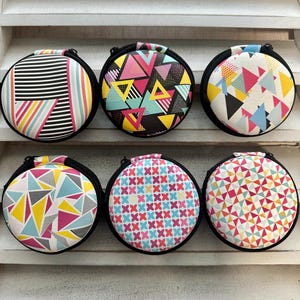 May include: Six round zippered cases with colorful geometric patterns. Designs include triangles, stripes, and cross patterns in various colors like pink, yellow, blue, and black. Each case has a black zipper and trim, and a small handle.