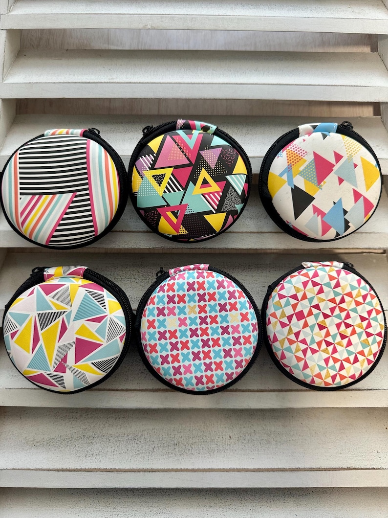 May include: Six round zippered cases with colorful geometric patterns. Designs include triangles, stripes, and cross patterns in various colors like pink, yellow, blue, and black. Each case has a black zipper and trim, and a small handle.