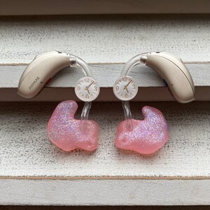 Puede incluir: Dos audífonos beige con tubo transparente y moldes de oído de color rosa brillante. Los audífonos tienen una pequeña esfera de reloj en el tubo con los números 9, 12, 3 y 6. El texto "PHONAK" es visible en uno de los audífonos.