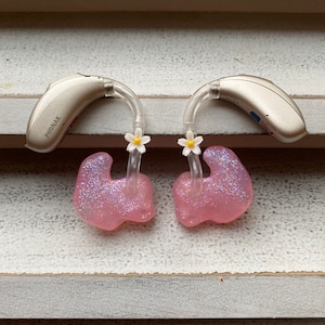 Könnte beinhalten: Zwei beige Hörgeräte mit individuellen rosa Glitzer-Ohrformen. Auf den Hörgeräten ist der Markenname "PHONAK" aufgedruckt. Jedes Hörgerät ist mit einer rosa Ohrform mit einem kleinen weißen Gänseblümchen-Akzent verbunden.
