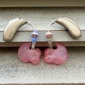 May include: Two beige Phonak hearing aids with clear tubing and pink glittery ear moulds. The tubing is decorated with small cartoon bunny figures. The hearing aids are resting on a white, weathered wooden surface.
