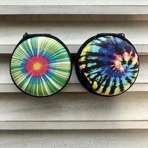May include: Two round, zippered cases with tie-dye designs. One case features a green, blue, and red spiral pattern, while the other displays a blue, yellow, and black tie-dye design. Both cases have a black border and zipper.