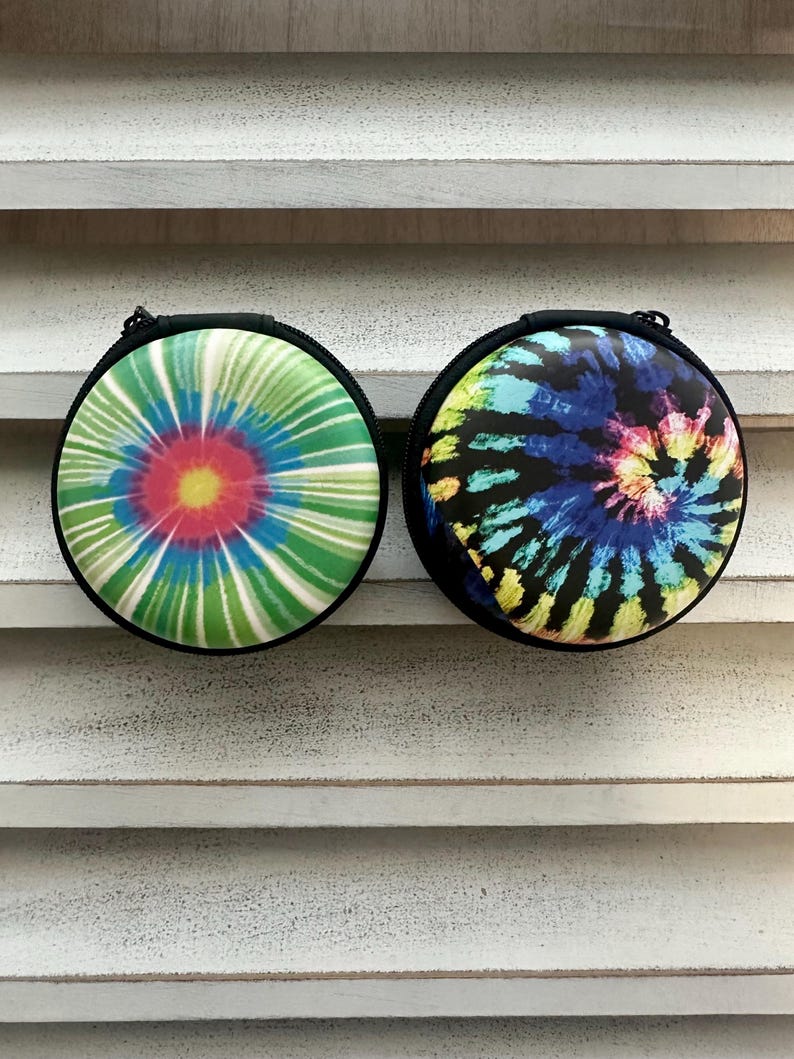 May include: Two round, zippered cases with tie-dye designs. One case features a green, blue, and red spiral pattern, while the other displays a blue, yellow, and black tie-dye design. Both cases have a black border and zipper.