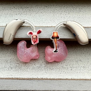 Könnte beinhalten: Zwei Phonak-Hörgeräte mit individuellen rosa Glitzer-Ohrpassstücken. Jedes Gerät hat einen dekorativen Aufkleber: einen mit einer Figur in einem Hasenkostüm, den anderen mit einer Beinlampe.