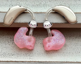 Puede incluir: Dos audífonos beige con tubo transparente y moldes de oído rosa. Los moldes de oído tienen brillo y un pequeño amuleto de perro de dibujos animados en cada tubo.