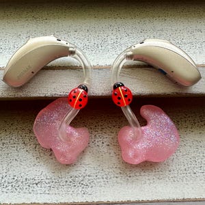 May include: A pair of beige hearing aids with pink ear moulds and ladybug charms. The hearing aids are labelled "PHONAK".