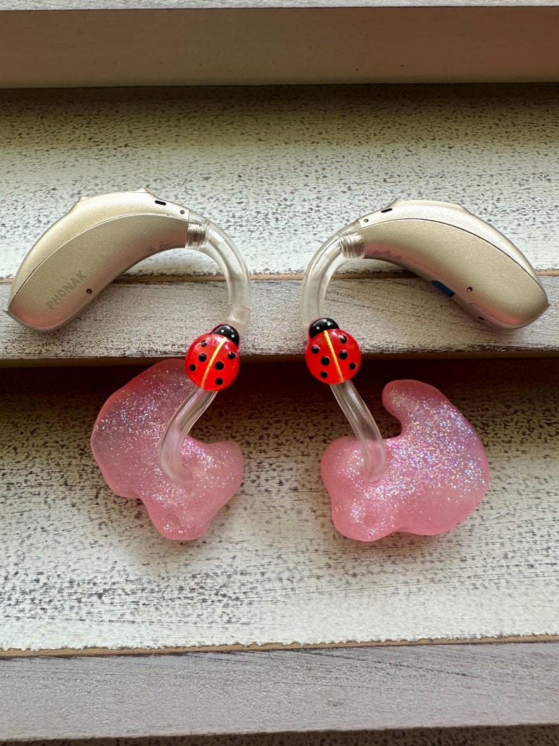 May include: A pair of beige hearing aids with pink ear moulds and ladybug charms. The hearing aids are labelled "PHONAK".