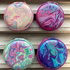 May include: Four round, zippered cases with a marbled design. The cases feature a variety of colors, including pink, purple, blue, and green. Each case has a different color zipper and trim, and a small handle.