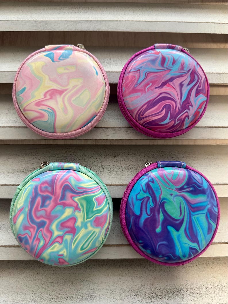 May include: Four round, zippered cases with a marbled design. The cases feature a variety of colors, including pink, purple, blue, and green. Each case has a different color zipper and trim, and a small handle.