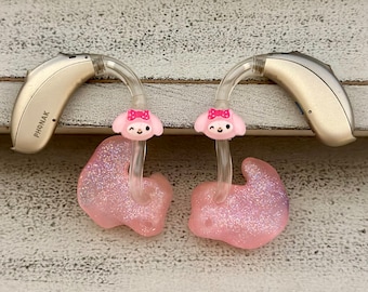 Puede incluir: Dos moldes de oído de color rosa brillante con un pequeño lazo rosa y una cara de animal de dibujos animados en cada uno. Los moldes de oído están unidos a audífonos beige con la etiqueta "PHONAK".