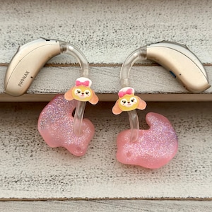 May include: Two pink sparkly hearing aid ear molds with a clear tube and a Disney character sticker on each. The stickers feature a cartoon dog with a pink bow and a yellow dress. The hearing aids are attached to a beige hearing aid device with the word "PHONAK" printed on the side.