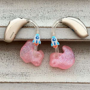 May include: Two beige hearing aids with clear tubing and pink sparkly ear moulds. The tubing has a small blue rocket ship charm attached to each side.