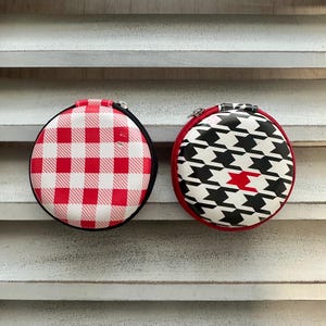 May include: Two round zippered cases. One case has a red and white gingham pattern, the other has a black and white houndstooth pattern with a red accent. Both cases have black trim and silver zippers.