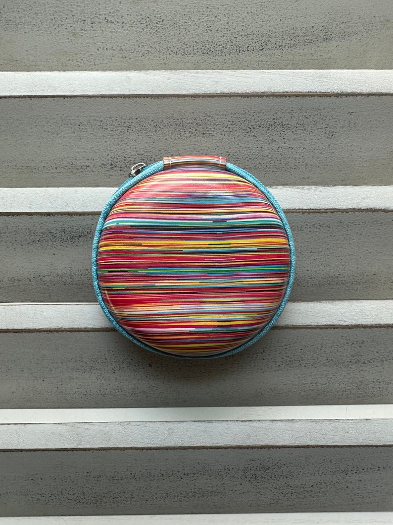 Peut inclure: Une pochette ovale de couleur ray&eacute;e avec une bordure bleue. Le sac a une fermeture &eacute;clair et une petite poign&eacute;e.