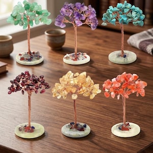 Op de afbeelding: Zes decoratieve boomsculpturen met stammen en takken van koperdraad, versierd met diverse getrommelde edelstenen in groen, paars, turkoois, donkerrood, geel en oranje, elk gemonteerd op een ronde stenen voet met kleine steentjes.