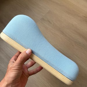 May include: A curved, ergonomic support cushion upholstered in a light blue fabric, featuring a smooth, natural-toned wooden base. The object has a distinct, elongated, wave-like silhouette with rounded edges and a textured textile surface.