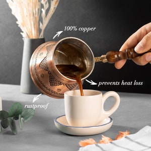 May include: A copper coffee pot with a wooden handle pouring coffee into a white cup. The pot has an intricate design and the text "100% copper" is written above it. The text "rustproof" is written below the pot and the text "prevents heat loss" is written to the right of the pot.