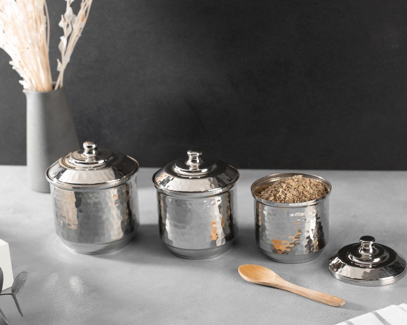 May include: Three silver hammered metal canisters with lids, one filled with brown grains, and a wooden spoon on a gray surface.