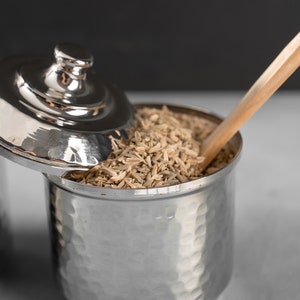 May include: A silver metal container with a lid, filled with fennel seeds. A wooden spoon rests on top of the seeds.