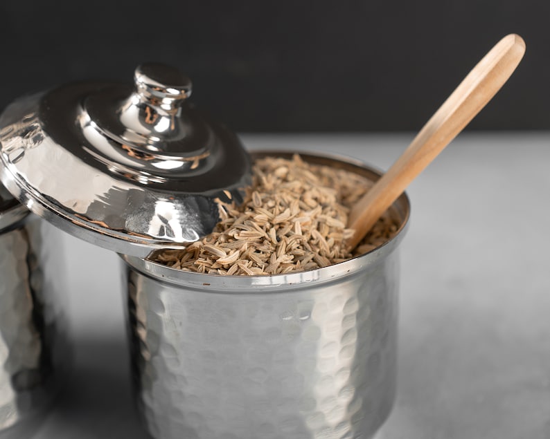 May include: A silver metal container with a lid, filled with fennel seeds. A wooden spoon rests on top of the seeds.