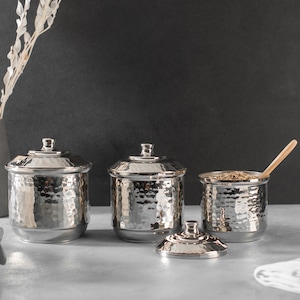 May include: Three silver hammered metal canisters with lids, one canister is open and contains dried herbs with a wooden spoon inside.