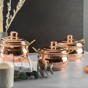 May include: Three copper pots with hammered texture and gold handles. The pots have lids and are arranged on a gray surface.