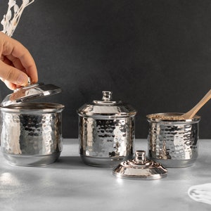 May include: Three silver hammered metal canisters with lids, one lid is open. The canisters are on a gray surface. One canister has a wooden spoon inside.