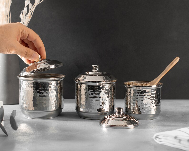 May include: Three silver hammered metal canisters with lids, one lid is open. The canisters are on a gray surface. One canister has a wooden spoon inside.