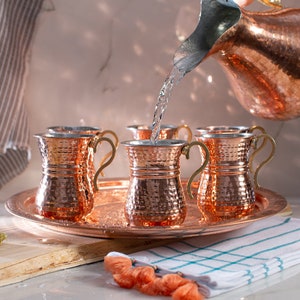 Op de afbeelding: Een set van vier gehamerde koperen bekers met gouden handvatten op een koperen dienblad. Een koperen kan giet water in een van de bekers.