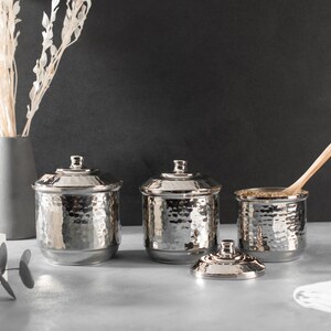 May include: Three silver hammered metal canisters with lids, one with a wooden spoon inside, on a gray countertop.