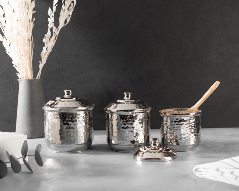 May include: Three silver hammered metal canisters with lids, one with a wooden spoon inside, on a gray countertop.
