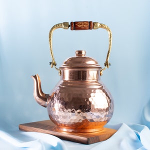 May include: A hammered copper teapot with a wooden handle and a gold-toned spout. The teapot is sitting on a wooden cutting board.