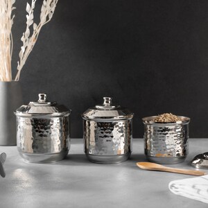 May include: Three silver hammered metal canisters with lids. The canisters are arranged in a row on a gray surface. A wooden spoon and a white and gray striped kitchen towel are in the foreground.