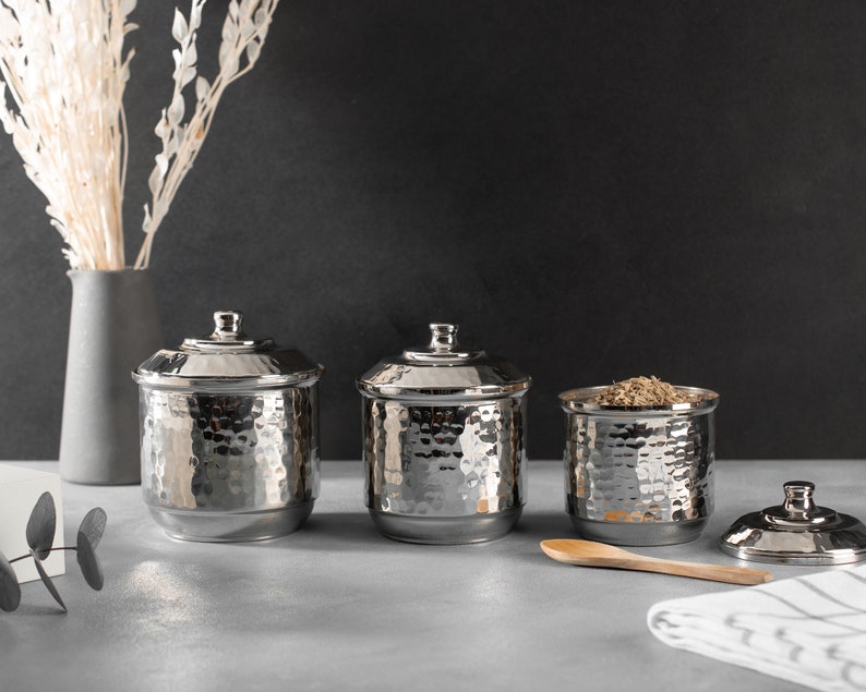 May include: Three silver hammered metal canisters with lids. The canisters are arranged in a row on a gray surface. A wooden spoon and a white and gray striped kitchen towel are in the foreground.