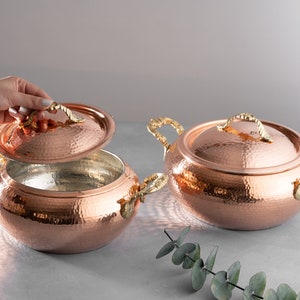 May include: Two copper pots with lids and brass handles. The pots have a hammered finish and are sitting on a gray surface.