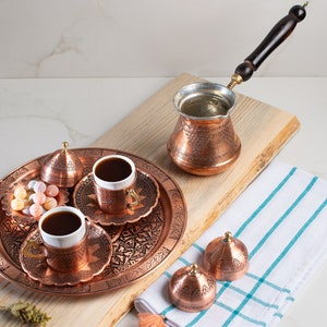May include: A copper serving tray with intricate designs holds two small copper coffee cups with saucers, a small bowl of colorful candies, and a lidded copper container. The tray is on a wooden surface next to a copper coffee pot with a wooden handle.