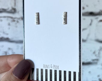May include: A pair of silver bar earrings with a textured surface. The earrings are displayed on a white card with black and white stripes and the text "Hunt & Peck".