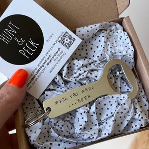 May include: A silver bottle opener with a key ring attached. The bottle opener has the text "WISH YOU WERE... BEER" engraved on it. The bottle opener is in a small box with white tissue paper with black polka dots.