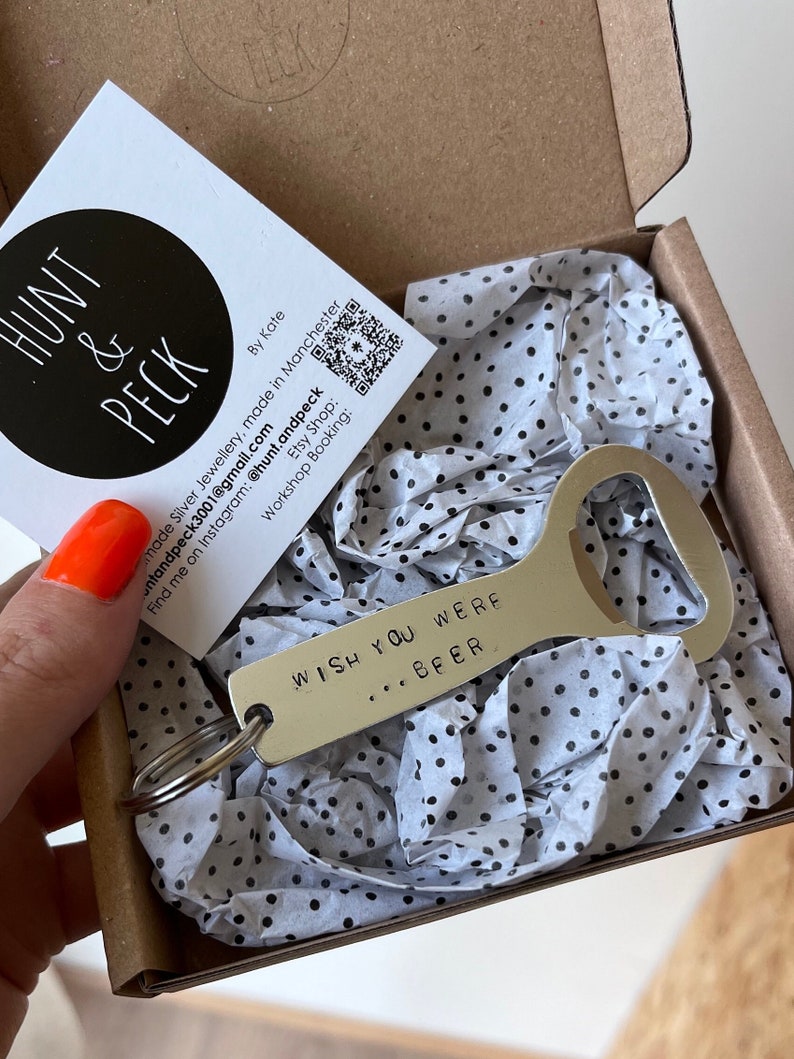 May include: A silver bottle opener with a key ring attached. The bottle opener has the text "WISH YOU WERE... BEER" engraved on it. The bottle opener is in a small box with white tissue paper with black polka dots.