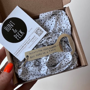 May include: A silver bottle opener keychain with the text "SURVIVING ADULTHOOD... ONE BEER AT A TIME" engraved on it. The keychain is packaged in a small white box with black polka dots.