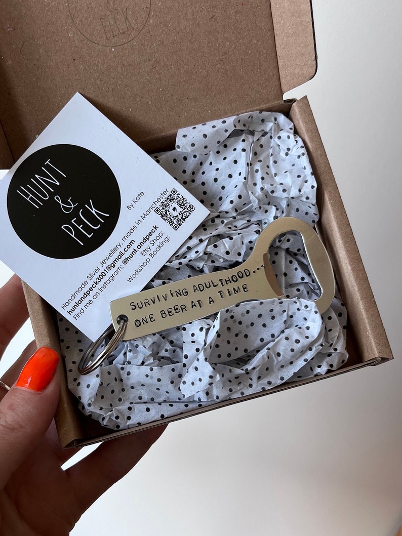 May include: A silver bottle opener keychain with the text "SURVIVING ADULTHOOD... ONE BEER AT A TIME" engraved on it. The keychain is packaged in a small white box with black polka dots.