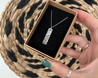 May include: A silver necklace with a rectangular pendant featuring heart designs is displayed in a small, open cardboard box with a black interior. The necklace is on a silver chain. The box is held against a woven basket.