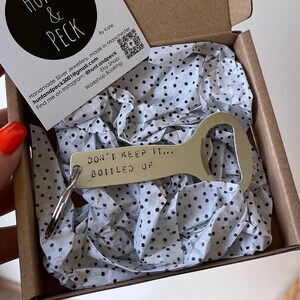 May include: A silver bottle opener keychain with the text "DON'T KEEP IT... BOTTLED UP" engraved on it. The keychain is in a small box with white tissue paper.
