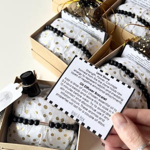 May include: A collection of lava stone bracelets, each with a silver-coloured tube, presented in small gift boxes. Each box includes a bracelet, a small bottle, and a card with text. The boxes are arranged on a white surface.