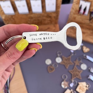 May include: A silver bottle opener keychain with the text "Save Water... Drink Beer" engraved on it. The keychain is held in a hand with yellow nail polish.
