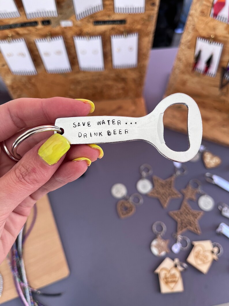 May include: A silver bottle opener keychain with the text "Save Water... Drink Beer" engraved on it. The keychain is held in a hand with yellow nail polish.