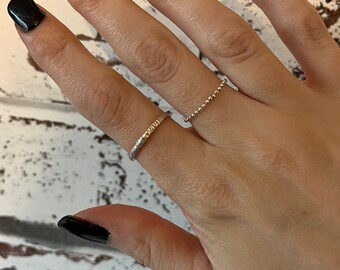 May include: Two silver rings on a hand with black nail polish. The ring on the index finger is a thin band with a textured surface. The ring on the middle finger is a delicate chain with small beads.
