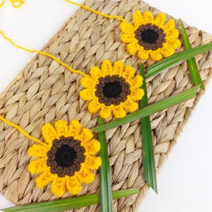 May include: Three crocheted sunflowers with brown centres and yellow petals are arranged on a woven straw mat with green leaves.